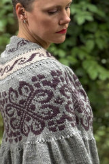 Side view of woman wearing knit shawl, looking over her right shoulder. The shawl is knit in pale grey with bands of colorwork in dark purple and pale peach.  thumbnail