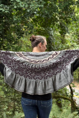 Back view of woman holding semi-circular shawl open. The top edge of the shawl is straight, and there are bands of filigree patterns on a grey background. The colorwork is knit in dark purple and pale peach.  thumbnail