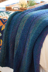 Knit blanket draped over the edge of a bed, with a wooden tray in the background. The blanket is knit in a color changing yarn in shades of blue, green, and purple. The throw also has horizontal textured stripes of stockinette and garter.  thumbnail