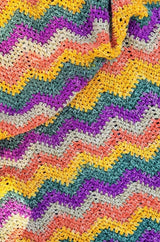 Top down view of crochet chevron blanket in self-striping yarn. The yarn is mustard yellow, green, purple, beige and orange.  thumbnail