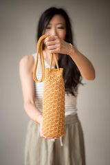 Woman holds a crochet water bottle holder toward camera, standing against a beige backdrop.  thumbnail
