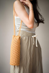 Side view of woman wearing water bottle holder over shoulder, with long strap. The top of the holder is at her hip. The bag is made with a solid textured fabric, in a warm camel color.  thumbnail