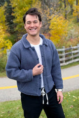 Man in a white shirt and dark pants stands outside with fall foliage and a white wooden fence behind. He also wears a knit jacket with a collar, button band, and pockets.  thumbnail