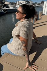 Side view of woman sitting on a dock in a short sleeve sweater. The sweater has ribbed edges and a textured body.  thumbnail