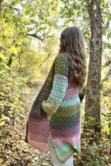 Side back view of woman standing in the woods. She wears a long crochet cardigan, with long sleeves, pockets, and a split hem.  thumbnail