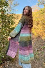 Side back view of woman in long crochet cardigan with her face turned toward camera. The cardigan is made in self-striping yarn in shades of green, blue, pink, and purple. The cardigan hangs to mid-calf and has a split hem up to the knee. The sleeves are long and there are also side pockets.  thumbnail