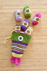 Fish pouch in greens and purples laid on bamboo surface, with small balls of yarn spilling from the opening.  thumbnail