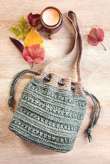 Crochet shoulder bag laid on pale wooden surface, with an unlit candle and fall leaves scattered around.  thumbnail