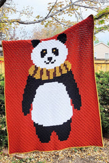 Crochet afghan hangs from a tree branch in a backyard. The afghan has a panda wearing a scarf, on a red-orange background.  thumbnail