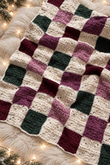 Crochet patchwork blanket laid on a white faux-fur rug. The blanket squares are cream, dark green, burgundy, dark pink, and medium pink.  thumbnail