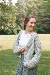 Woman in a crochet cardigan standing in a grassy backyard. The cardigan has a mesh texture and solid cuffs and buttonband. It's made in a pale green and worn over a white top and floral print skirt.  thumbnail