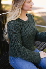 Side view of woman wearing green crochet pullover with long sleeves, sitting on a bench.  thumbnail