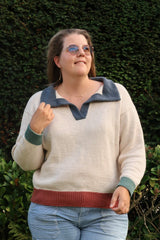 Woman in a knit polo sweater standing in front of a green hedge. The sweater is mainly cream, with a terracotta hem, sage green cuffs, and a denim blue collar.  thumbnail