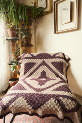 Crochet lapghan draped over a chair in a a room corner, with plants and framed photos on the wall.  thumbnail