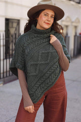 Woman in a knit cabled poncho, felt wide brimmed hat, and rust colored pants stands on a city sidewalk.  thumbnail