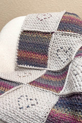 Crochet patchwork blanket laid on white armchair. The squares are a parchment color with embroidered hearts, alternating with squares in a self-striping yarn.  thumbnail