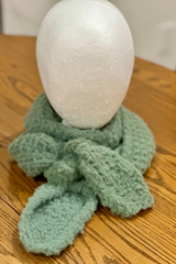 sage green knitted wrap scarf wrapped loosely around a white foam mannequin head resting on a wooden surface thumbnail