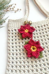 two crocheted red poinsettia appliqués with yellow centers and green leaves attached to a cream-colored textured crochet bag with wooden buttons thumbnail