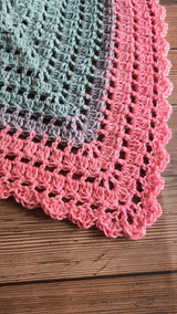 front view of a multicolored pastel pink, grey, and blue gradient crocheted triangle shawl with a lacy border, displayed over a beige fleece jacket thumbnail