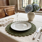 sage green crocheted round placemat with openwork lattice and scalloped edge set on a beige tablecloth with a white dinner plate, silverware, and a ceramic vase of hydrangeas thumbnail