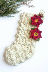two crocheted red poinsettia appliqués with yellow centers and green leaves attached to a cream-colored textured knit Christmas stocking hung against a white background thumbnail