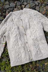 Flat lay of cabled knit cardigan on stones. The cardigan is a raglan style with long sleeves and two patch pockets on the front.  thumbnail