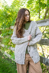 Woman standing next to wooden fence, with greenery in the background. She wears an olive top and tan pants, with an oatmeal cable knit cardigan. thumbnail