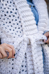 closeup of a person tying the waist belt on a cream-colored V-stitch crocheted openwork cardigan over a denim shirt thumbnail