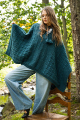 front view of a person wearing a teal-green knitted hooded poncho with an allover texture pattern and fringe tassel ties, paired with light wash denim jeans outdoors thumbnail