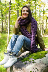 person wearing a violet openwork mesh crocheted scarf, olive jacket, and jeans against a wooded outdoor background thumbnail