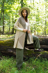 full-length view of a person wearing a cream granny square crocheted wrap, white shirt, olive pants, and a hat outdoors in a wooded setting. thumbnail