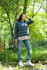 full-length view of a person wearing a two-tone blue striped knit pullover, striped shirt, jeans, and white boots outdoors in a wooded setting thumbnail