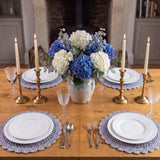 lavender crocheted round placemats set on a wooden dining table with place settings, brass candlesticks, and a blue and white hydrangea centerpiece thumbnail