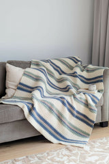striped crocheted blanket with blue, sage green, and beige stripes draped over a grey sofa with a white pillow and coffee mug beside it thumbnail