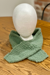 sage green knitted wrap scarf wrapped loosely around a white foam mannequin head resting on a wooden surface thumbnail