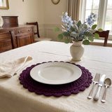 plum crocheted round placemat with openwork lattice and scalloped edge set on a beige tablecloth with a white dinner plate, silverware, and a vase of flowers thumbnail