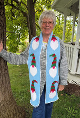 Woman standing in a green backyard, with a white wooden porch railing behind. She wears a crochet scarf in pale blue with Christmas gnomes.  thumbnail