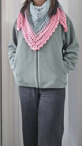 front view of a person wearing a pink crocheted triangle scarf with a lacy border over a grey fleece zip-up jacket and dark trousers thumbnail