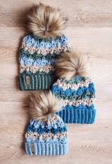 three handmade striped crochet beanies with faux fur pom-poms and ribbed cuffs arranged on a light wooden surface thumbnail