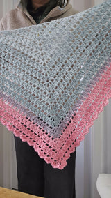 front view of a person wearing a multicolored pastel pink, grey, and blue gradient crocheted triangle shawl with a lacy border, displayed over a beige fleece jacket thumbnail