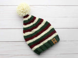 striped elf stocking cap with red, white, and green stripes, a white pom-pom, and a branded tag on a light wooden background thumbnail