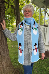 Woman standing in a green backyard, with a white porch railing behind. She has a crochet scarf with a pale blue background and snowmen along the length. The snowmen have black top hats and either red or green scarves. The scarf is wrapped once around her neck and hangs to mid-thigh.  thumbnail