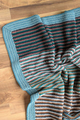 flat lay of a striped crochet baby blanket with a wide blue border on a wooden floor thumbnail